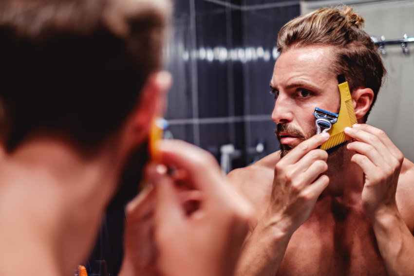 man shaving his beard with a beard shaping tool in a mirror and using a cartridge razor