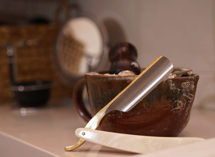 straight razor leaning on a shaving scuttle with brush and lather inside